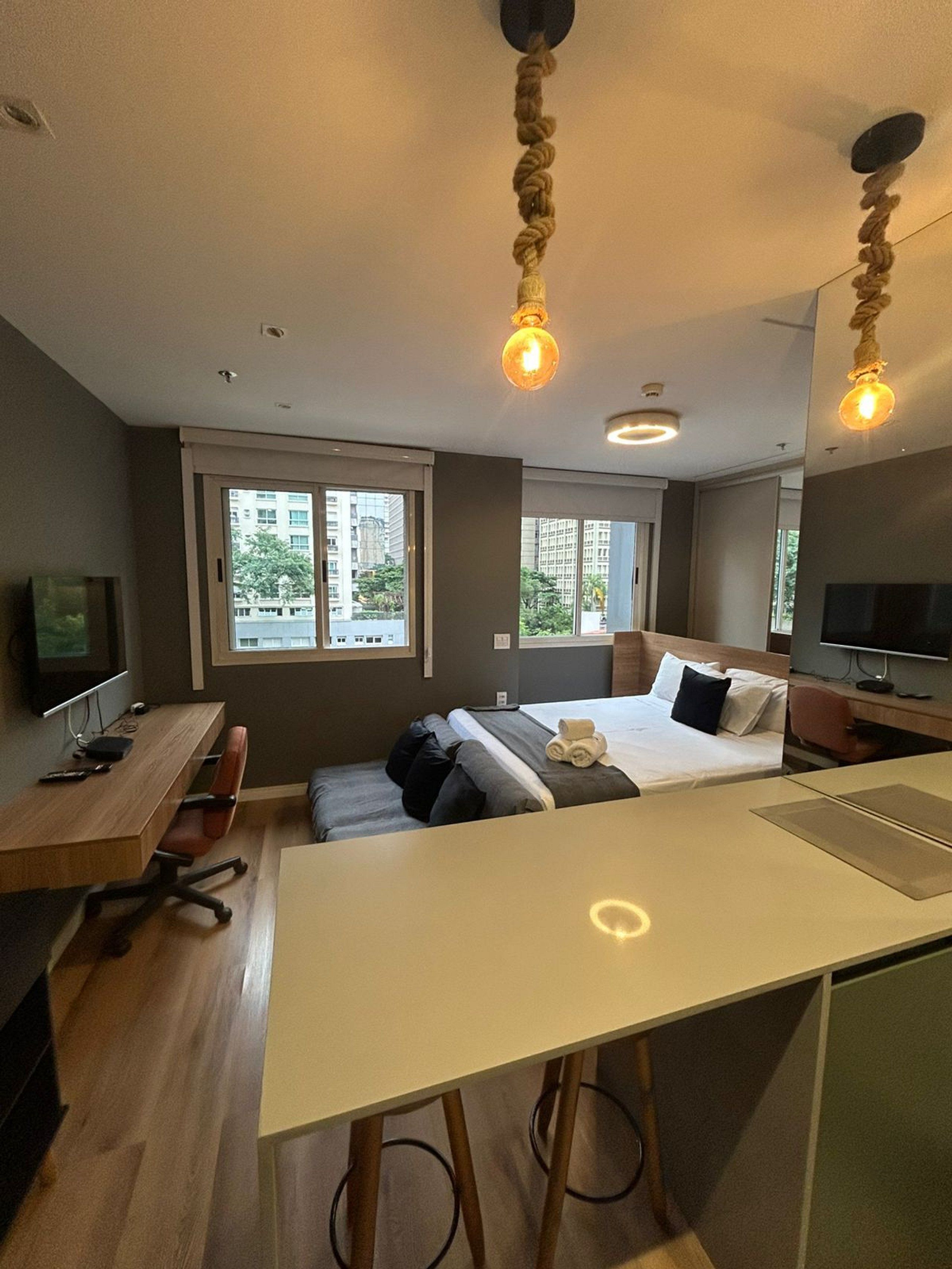 A modern hotel room with a bed, desk, and seating area. There are windows providing a view of the city.