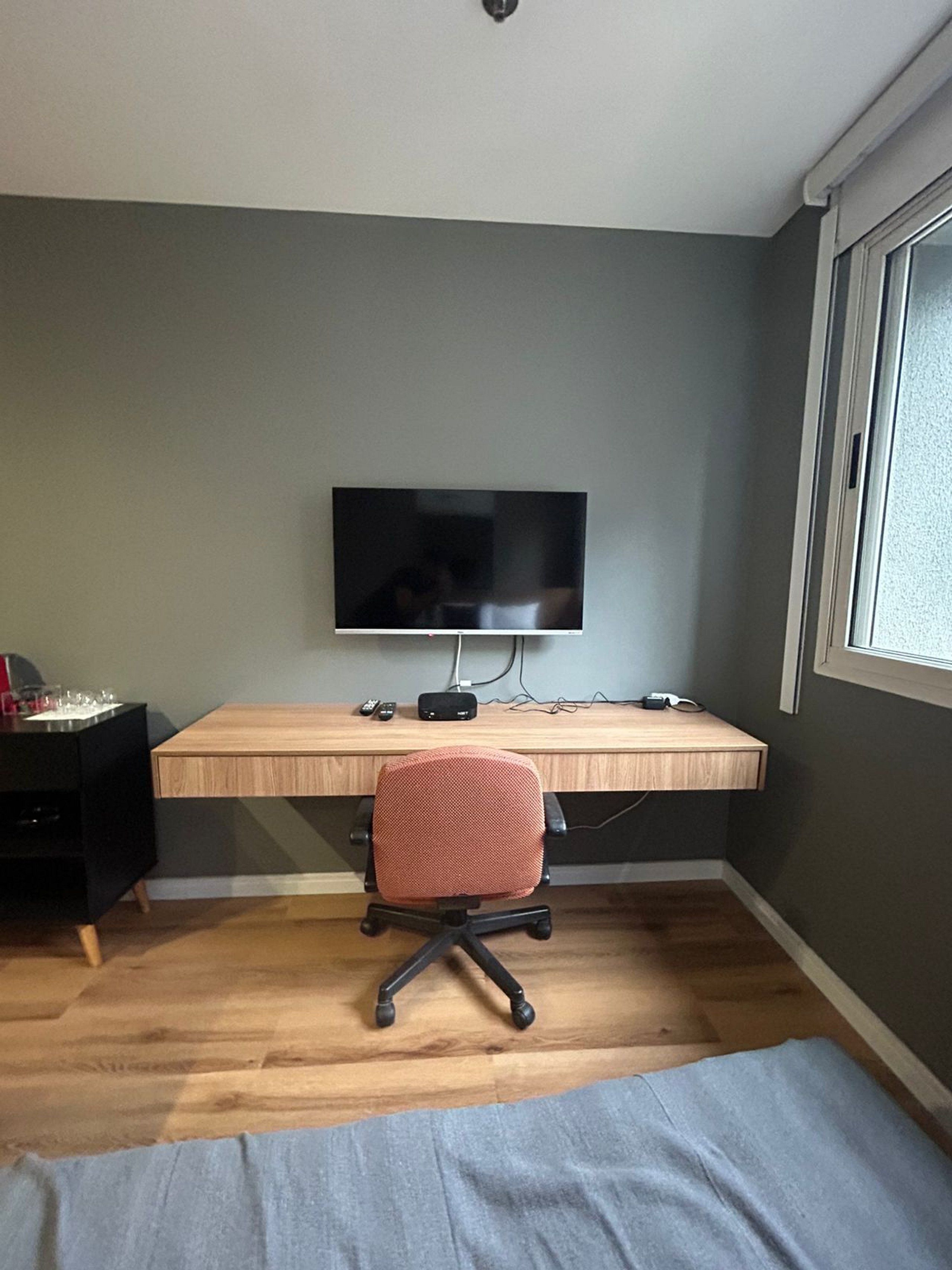 Une pièce avec un bureau en bois, un téléviseur fixé au mur, un fauteuil de bureau rouge et une fenêtre sur le côté droit.
