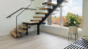 Una escalera moderna con barandillas de vidrio y peldaños de madera, que conduce a una habitación con un ventanal y vistas a árboles.