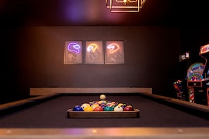 Een pooltafel met ballen, drie neonkunstwerken in lijsten en speelautomaten in een schemerige ruimte.