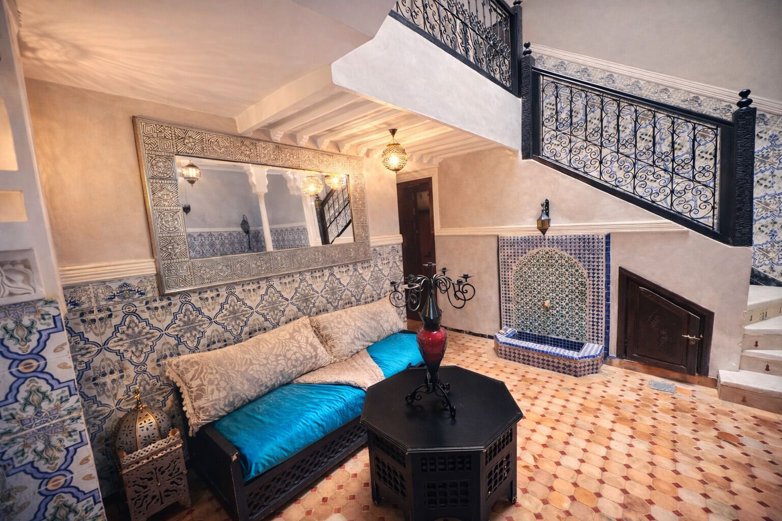 A room with a patterned floor, a sofa with cushions, a decorative lamp, a wall-mounted mirror, and a staircase with a patterned railing.