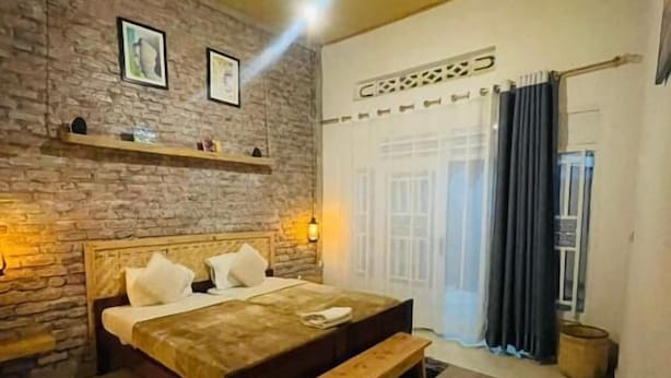 A bedroom with a bed, a bench, a rug, a brick wall, and a window with curtains. - Saish Stay (Muhoza)