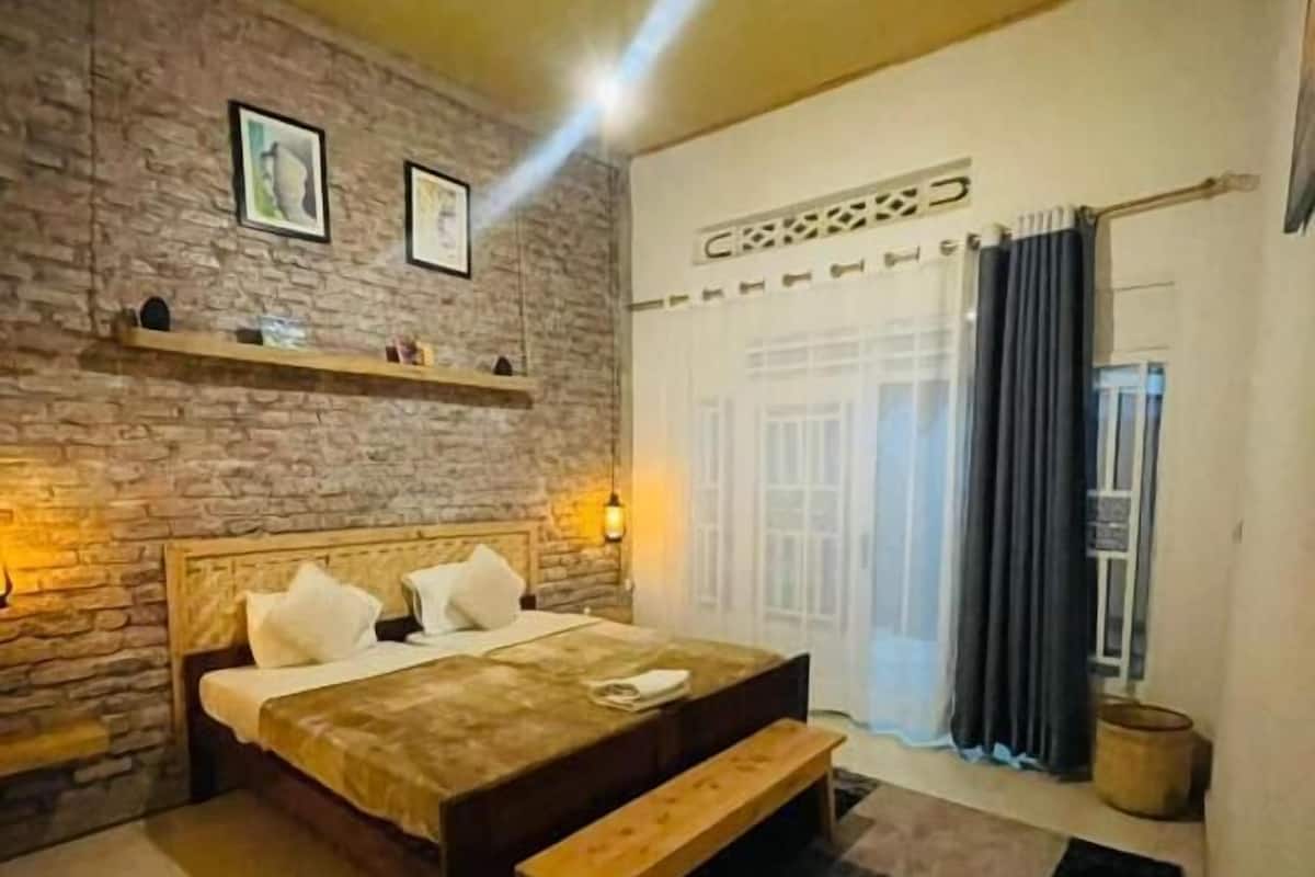 A bedroom with a bed, a bench, a rug, a brick wall, and a window with curtains.