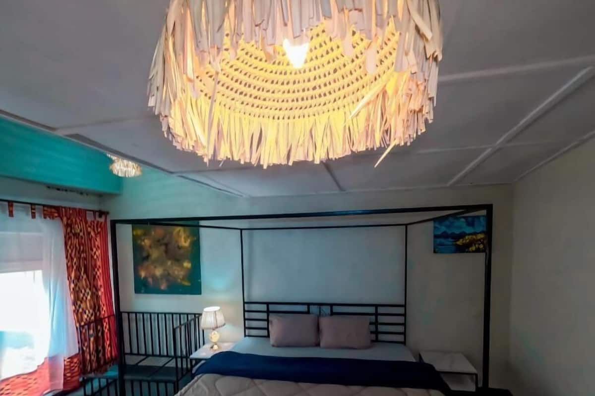 A bedroom with a four-poster bed, a hanging light fixture, a painting on the wall, and a window with curtains.