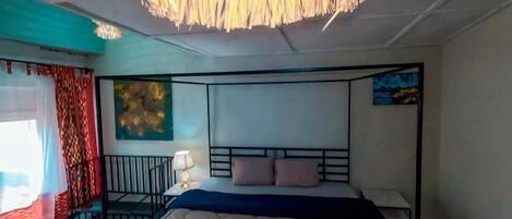 A bedroom with a four-poster bed, a hanging light fixture, a painting on the wall, and a window with curtains.
