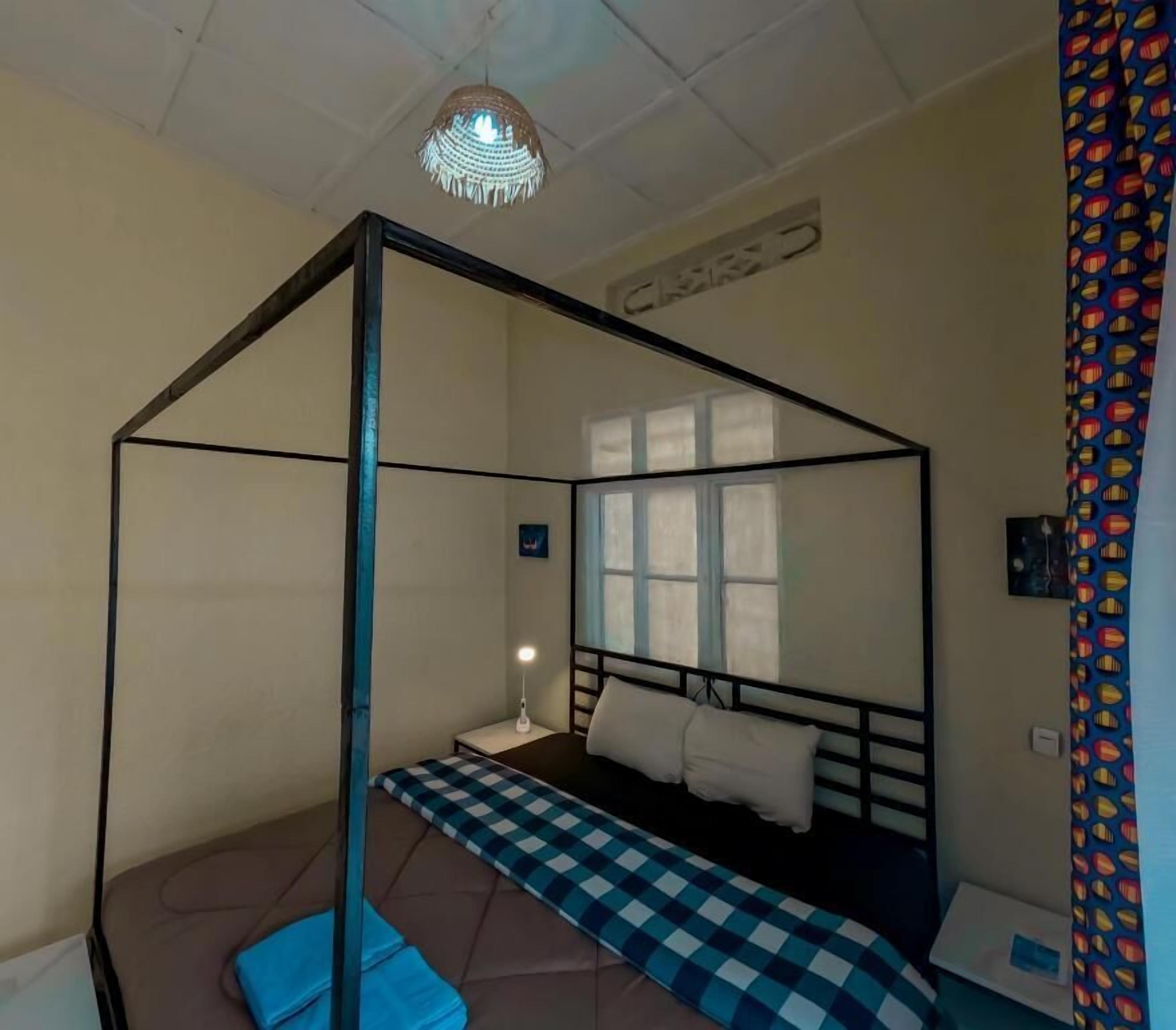 A room with a four-poster bed, a single bedside table with a lamp, a window with blinds, and a patterned curtain.