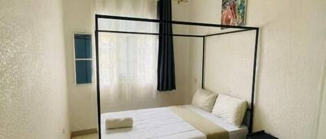 Een eenpersoonsbed met hoofdeinde, een nachtkastje, een klein raam met gordijnen en een schilderij aan de muur.