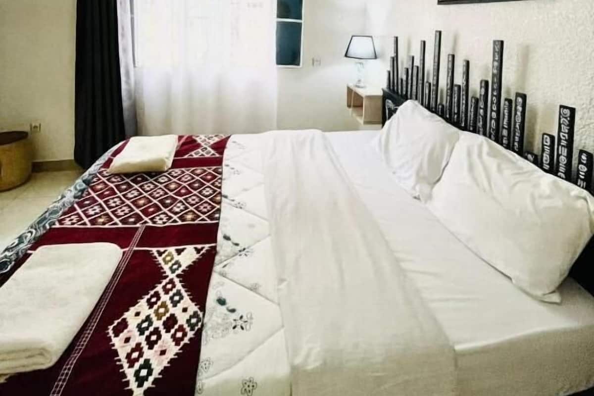 A hotel room with a bed, a desk, a chair, a window with curtains, and a painting on the wall.