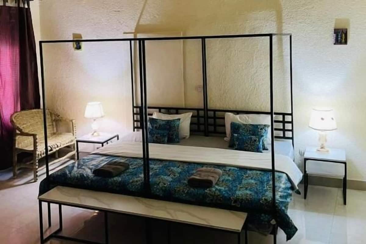 A room with a four-poster bed, a bench, two bedside tables with lamps, a chair, and a small picture on the wall.