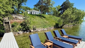 A wooden deck with lounge chairs and a small table, overlooking a river and a house in the background. - Delightful 6-bedroom cottage on charming Lake Carlos in Alexandria, MN (Alexandria)