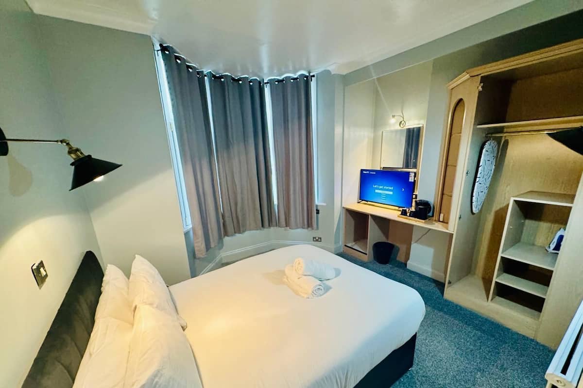 Quarto de hotel com cama, escrivaninha com computador, armário e janela com cortinas.