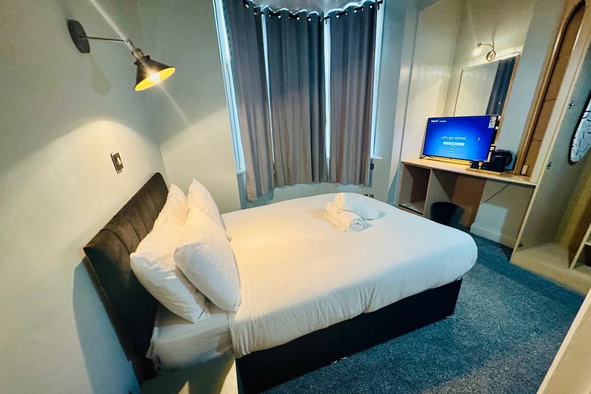 Quarto de hotel com cama, escrivaninha com computador, espelho e janela com cortinas.