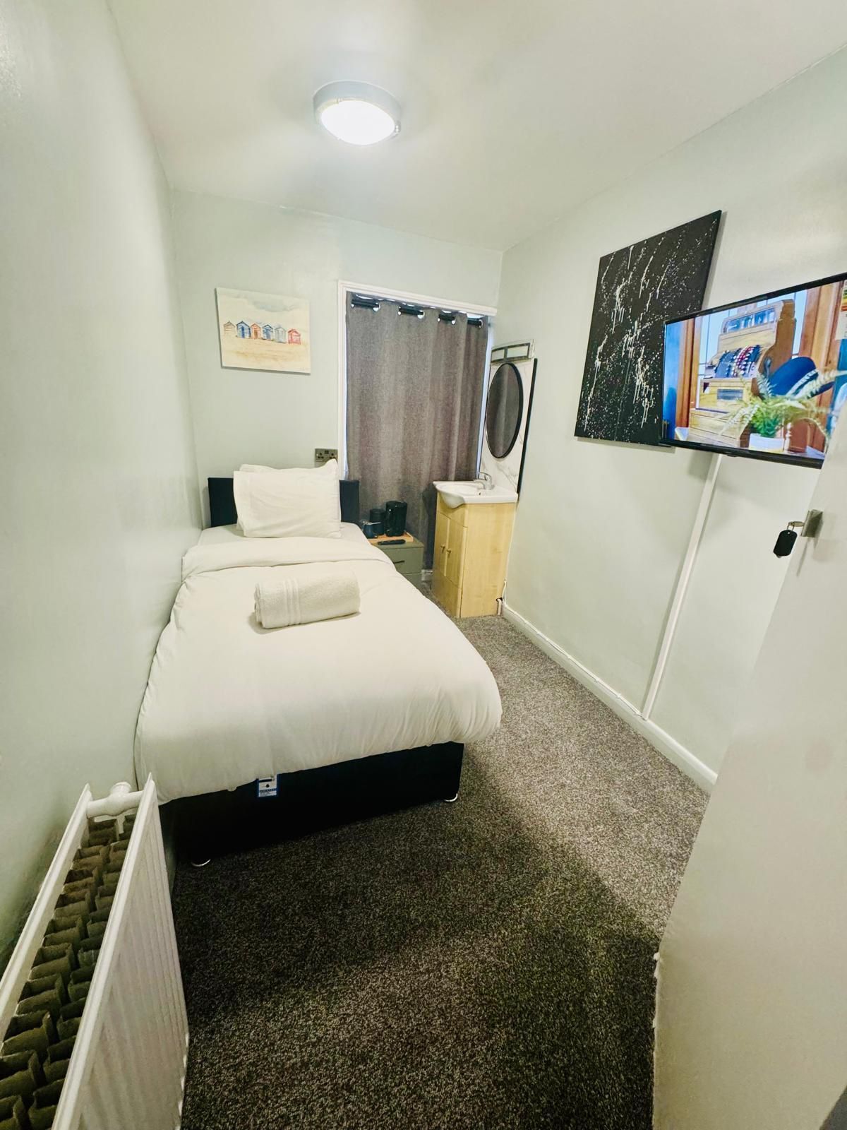 A hotel room with a bed, a small bedside table, a television, a painting, and a radiator.