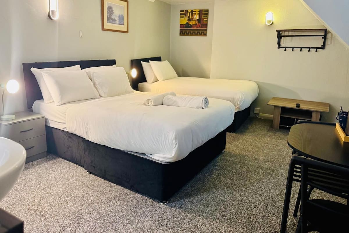 Quarto com duas camas, uma mesinha de cabeceira com abajur, um pequeno banco de madeira e uma escrivaninha com cadeira.