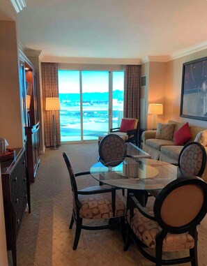 Ein Hotelzimmer mit einem Glastisch, Stühlen, einer Couch, einem Fernseher und einem großen Fenster mit Blick auf den Strand.
