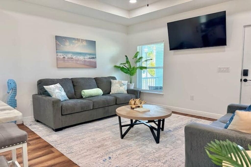 Ein Wohnzimmer mit grauem Sofa, einem runden Couchtisch, einem an der Wand befestigten Fernseher und einem Fenster mit Jalousien.