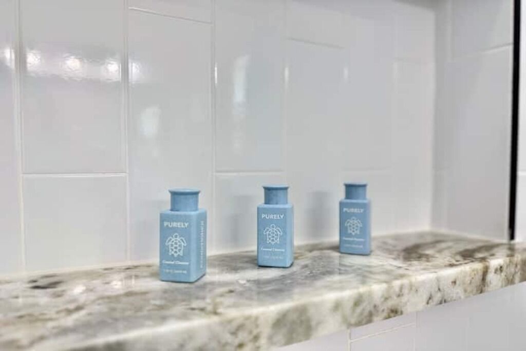 Un baño con azulejos blancos y tres botellas azules de champú Purely sobre una encimera de mármol.