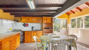 A kitchen with wooden cabinets, a dining table with chairs, and a view through large windows. - Holiday Home 'Jardim do Sol' with Sea View, Private Garden and Wi-Fi (Lugares)