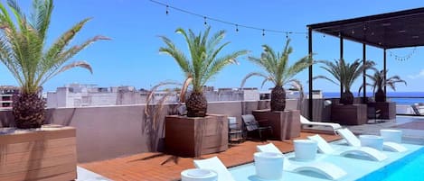 A rooftop pool area with lounge chairs, palm trees, and a clear view of the ocean.