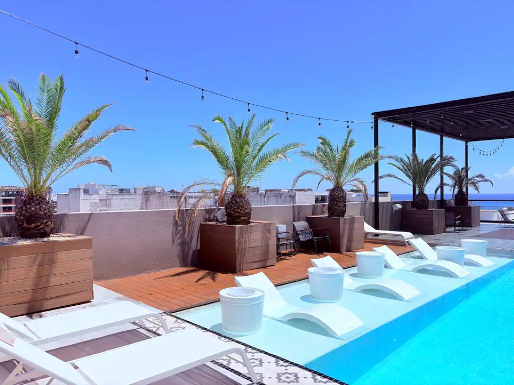A rooftop pool area with lounge chairs, palm trees, and a clear view of the ocean.