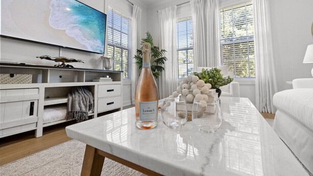 Una sala de estar moderna con un televisor de pantalla plana grande, una mesa de centro de mármol con objetos decorativos, muebles blancos y ventanas amplias con cortinas.