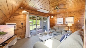 En mysig stuga med inredning som inkluderar en tv, soffa, soffbord, takfläkt och en skjutdörr i glas som leder ut till en uteplats.