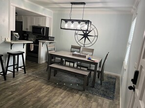 Uma sala de jantar com mesa de madeira, cadeiras, um lustre e uma cozinha ao fundo.