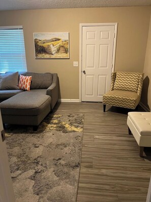 Una sala de estar con un sofá seccional gris, un sillón estampado, una puerta blanca y un cuadro en la pared.