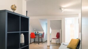 A room with a black bookshelf, a grey sofa with a yellow pillow, a dining table with red chairs, and a globe on top of the bookshelf. - LU Mercury lll - Old Town HITrental Apartment by Interhome (Lucerne)