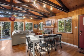 A wooden interior with a dining table, chairs, a fireplace, and large windows.