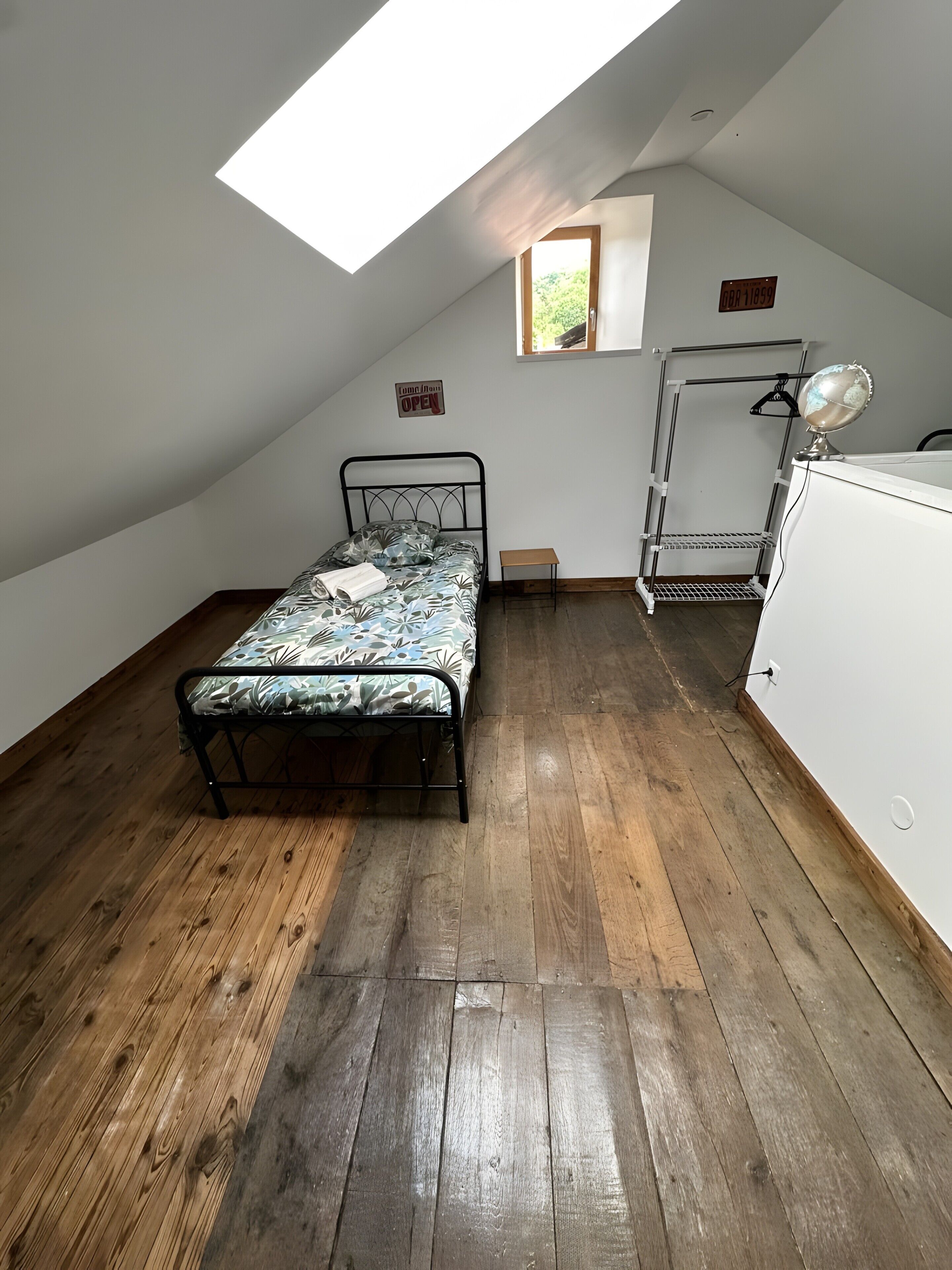 A room with a sloped ceiling, a bed with a patterned bedspread, a small table, a hanging rack, and a window with a view of greenery.