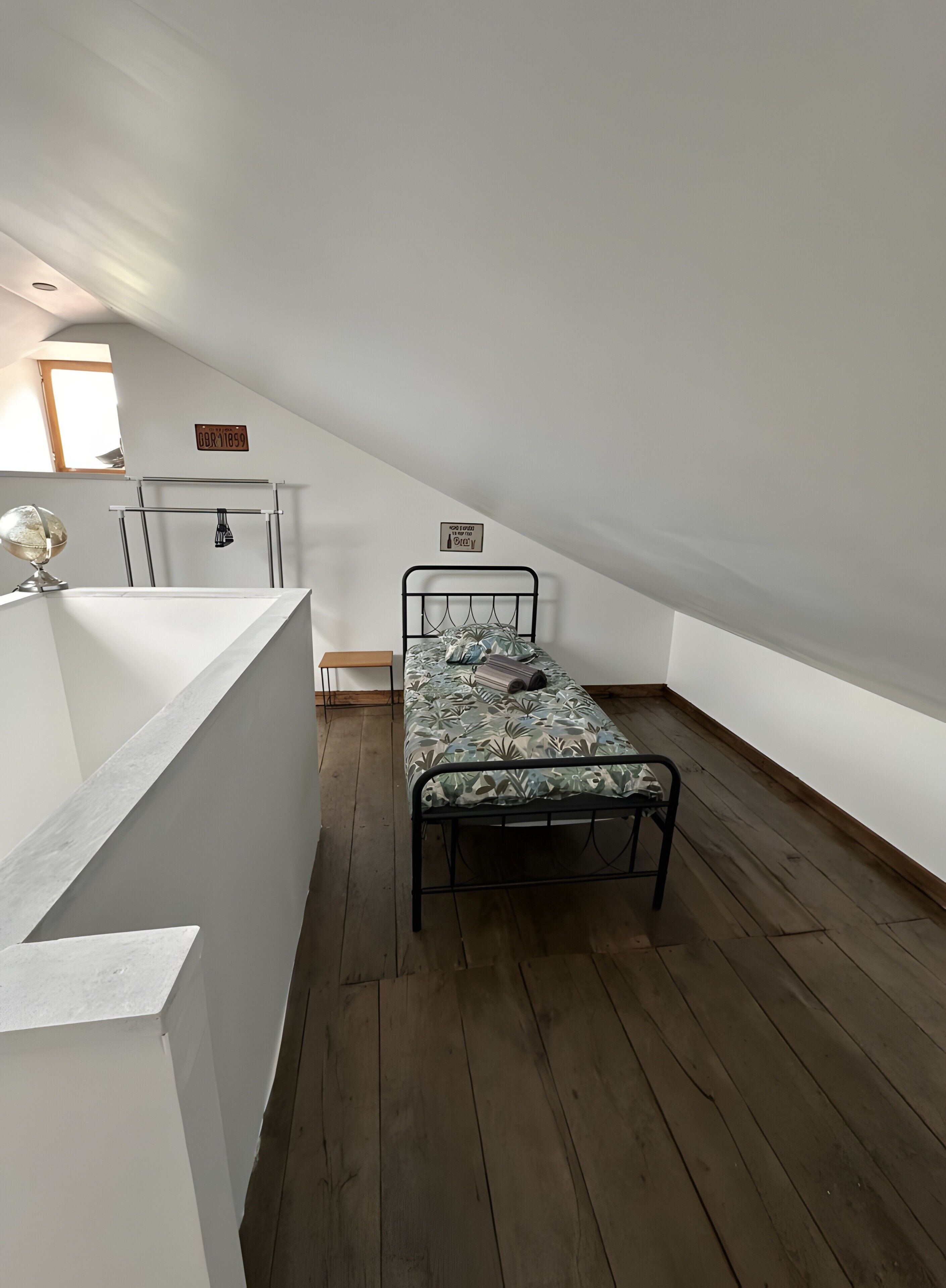 A small, single-bed room with a sloped ceiling, a wooden floor, a white wall, and a small window.