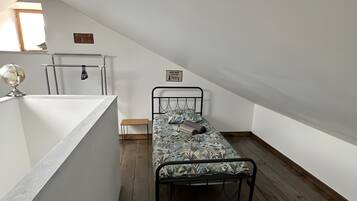 A small, single-bed room with a sloped ceiling, a wooden floor, a white wall, and a small window.