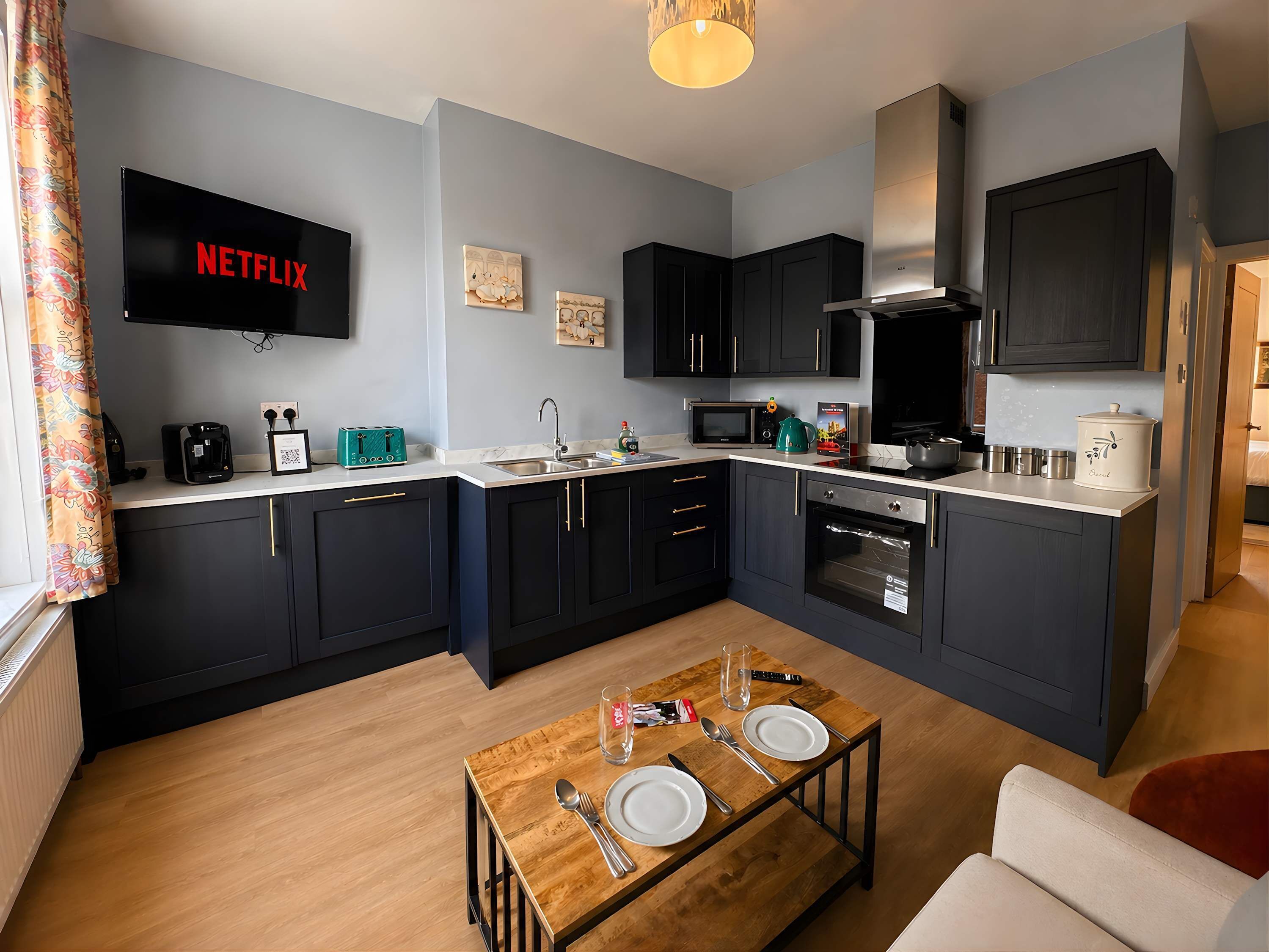A compact kitchen with a built-in oven, microwave, and coffee maker. A dining table with cutlery is set in the center.