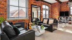 A living room with brick walls, a black leather sofa, a glass coffee table, and a television. - Modern Studio Flat York City Centre near Shambles (York)