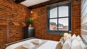 A bedroom with a brick wall, a bed with white linens, a dresser, a plant, and a window with blinds. - Modern Studio Flat York City Centre near Shambles (York)