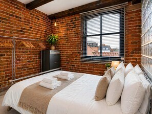 Un dormitorio con pared de ladrillo, cama con ropa de cama blanca, cómoda, planta y ventana con persianas.