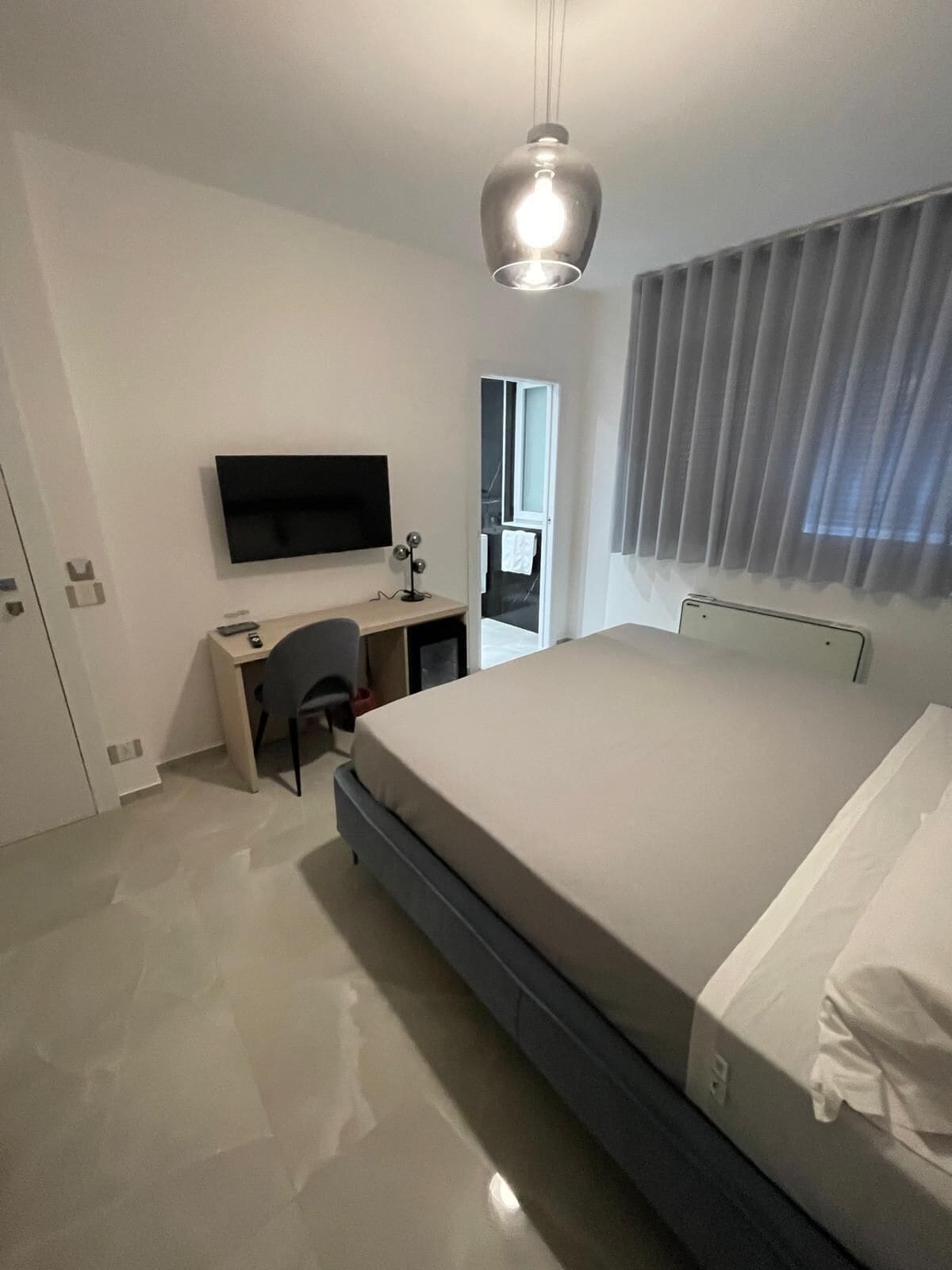 Un dormitorio moderno con cama, escritorio, silla, televisor y ventana con cortinas.