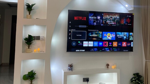 Un soggiorno moderno con televisore a parete che mostra diverse piattaforme di streaming, una ventola a soffitto ed elementi a incasso con piante decorative.