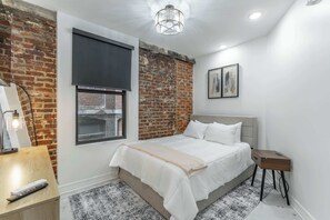 A bedroom with a bed, a nightstand, a lamp, a window, and a brick wall.