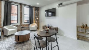 Una sala moderna con una mesa de centro redonda, dos sillones blancos, un sofá, un televisor montado en la pared y una mesita auxiliar con objetos decorativos.