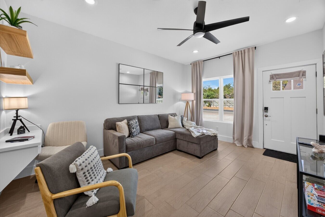 Un salon moderne avec un canapé d’angle gris, une chaise en bois, un bureau avec une lampe et un ventilateur de plafond.
