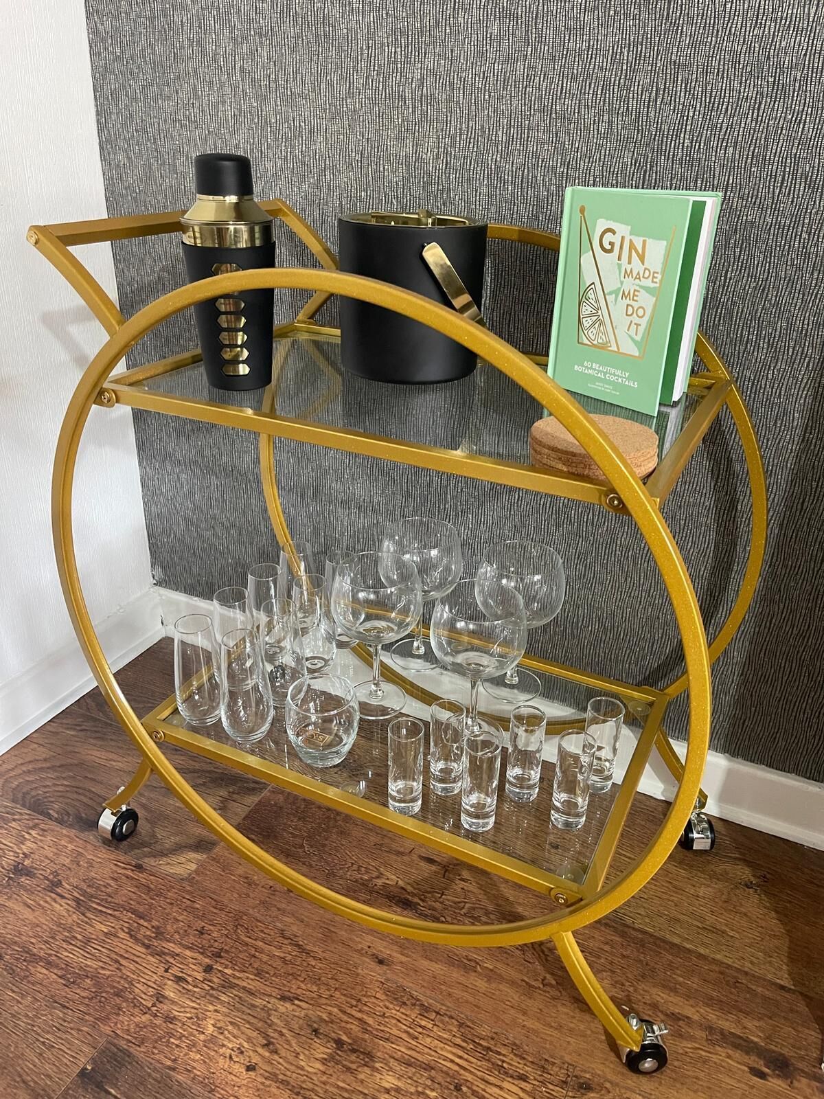 Ein goldfarbener Drahtkorb mit mehreren Gläsern und Flaschen, ein Buch mit dem Titel 'GIN MADE ME DO IT' und eine strukturierte graue Wand im Hintergrund.
