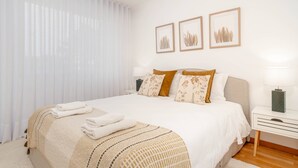 Un lit bien fait, recouvert d’une couvre-lit beige et blanche, avec deux oreillers à motifs floraux et une table de chevet surmontée d’une lampe.