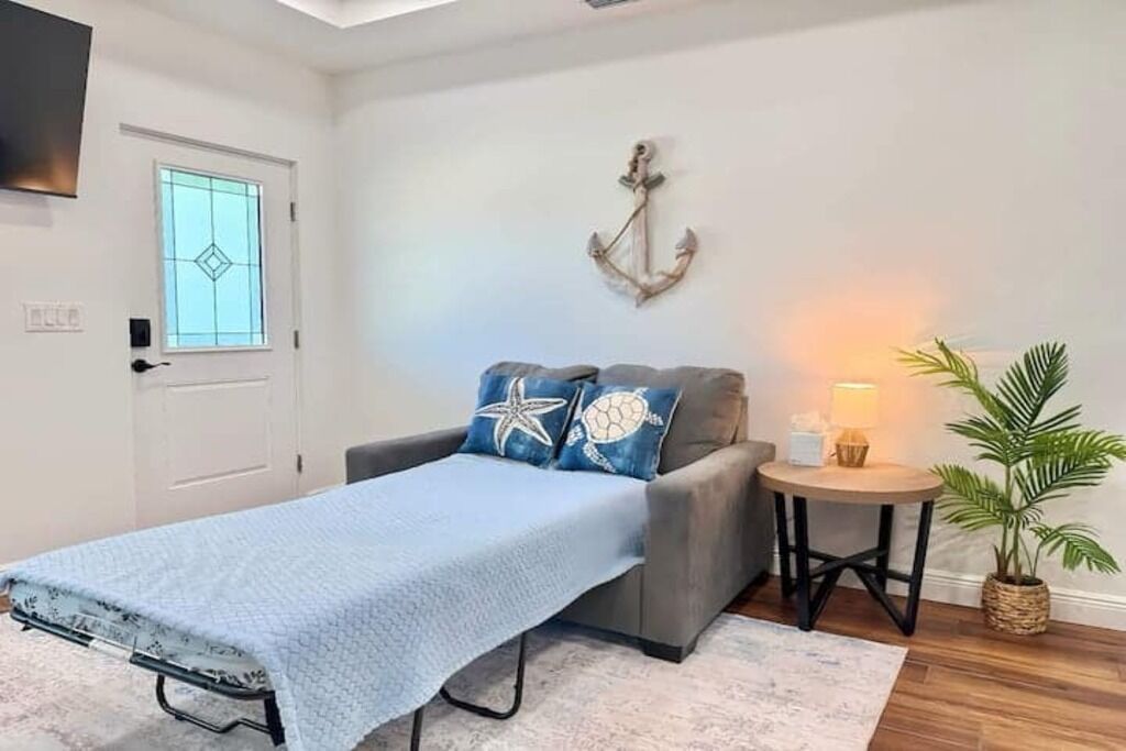 Una habitación con una cama, un sofá gris, una pequeña mesa redonda con una lámpara, una planta en maceta y una decoración de ancla en la pared.