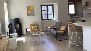 En moderne opholdsstue med sofa, lænestole, et lille rundt bord, et fjernsyn og et tekøkken med et vindue.