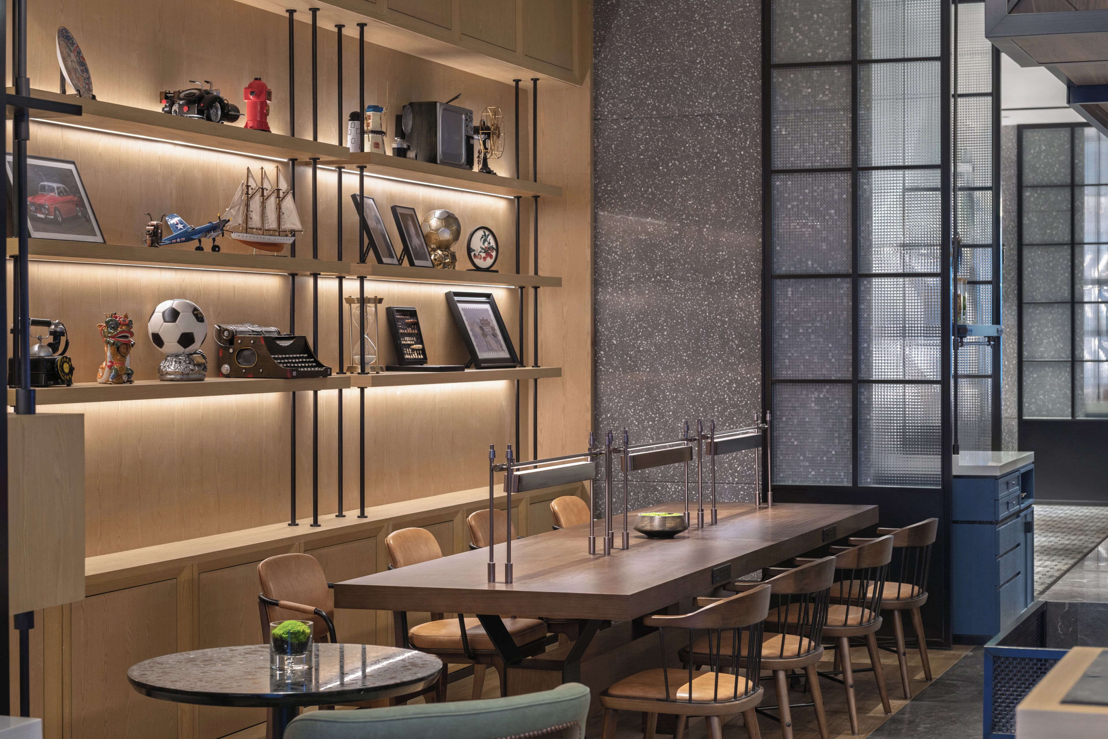 Un espace repas moderne avec une longue table et des chaises, un coin bar avec des étagères remplies d’objets, et une grande fenêtre offrant une vue dégagée.