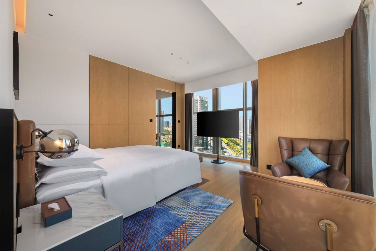 Quarto de hotel moderno com uma cama grande, uma televisão de tela plana, uma poltrona de couro e vista para a cidade por grandes janelas.