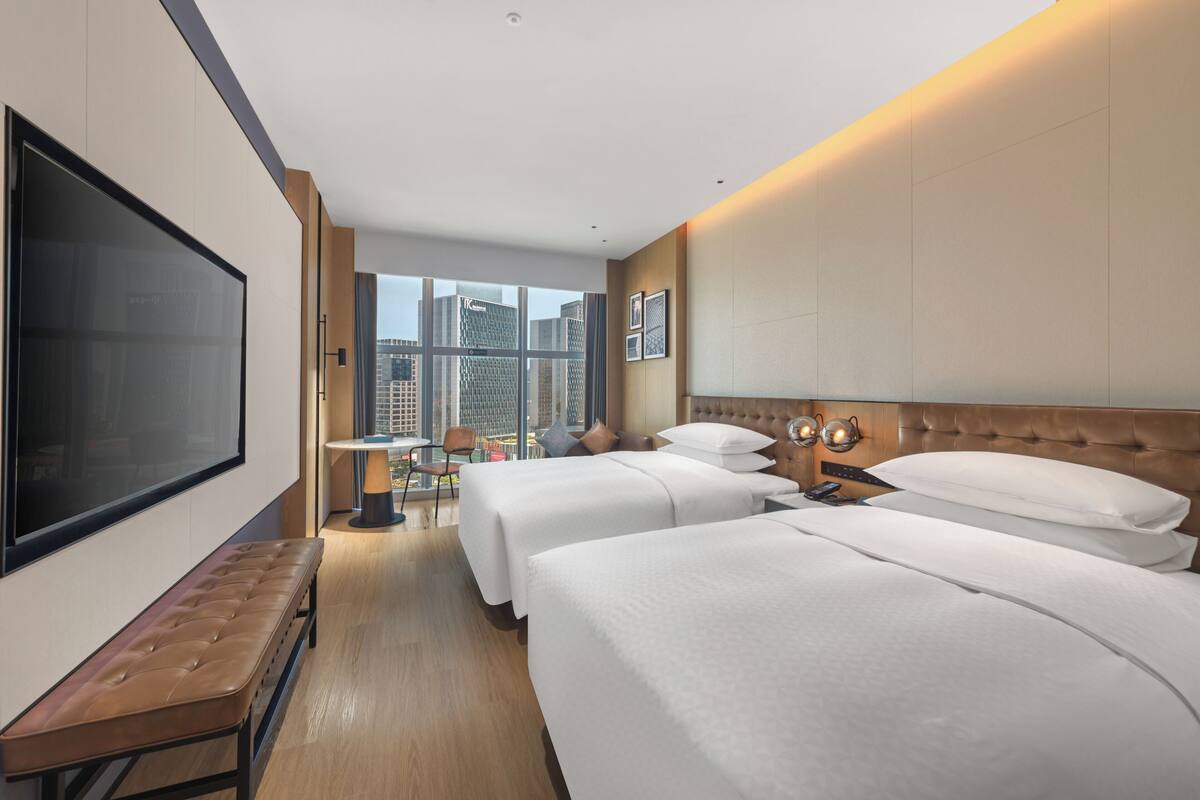 Quarto de hotel com duas camas, uma TV grande, um banco, uma mesa pequena e vista para a cidade.