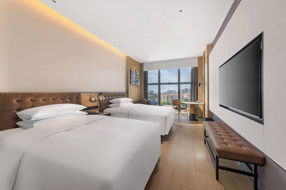 Quarto de hotel com duas camas, uma TV grande, um banco e vista da cidade.